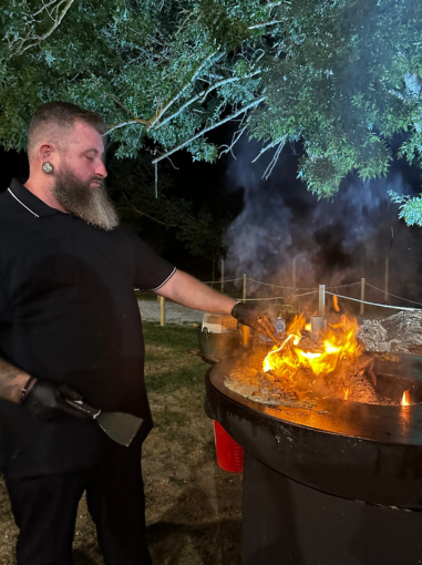 Chef Barbecue Man en action, cuisinant sur un feu ouvert lors d'un événement nocturne.